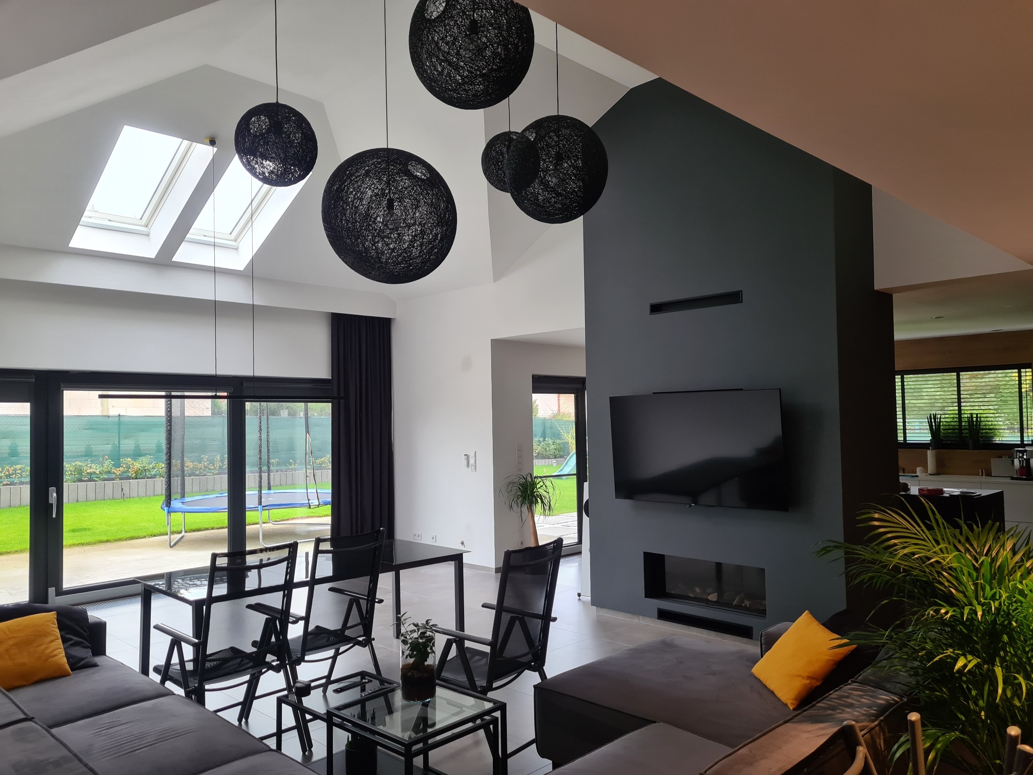 Nowoczesny salon z wysokim sufitem, oświetlony wiszącymi kulami z czarnego sznurka, szary narożnik z żółtą poduszką, widok na zielony ogród przez przeszklone drzwi.