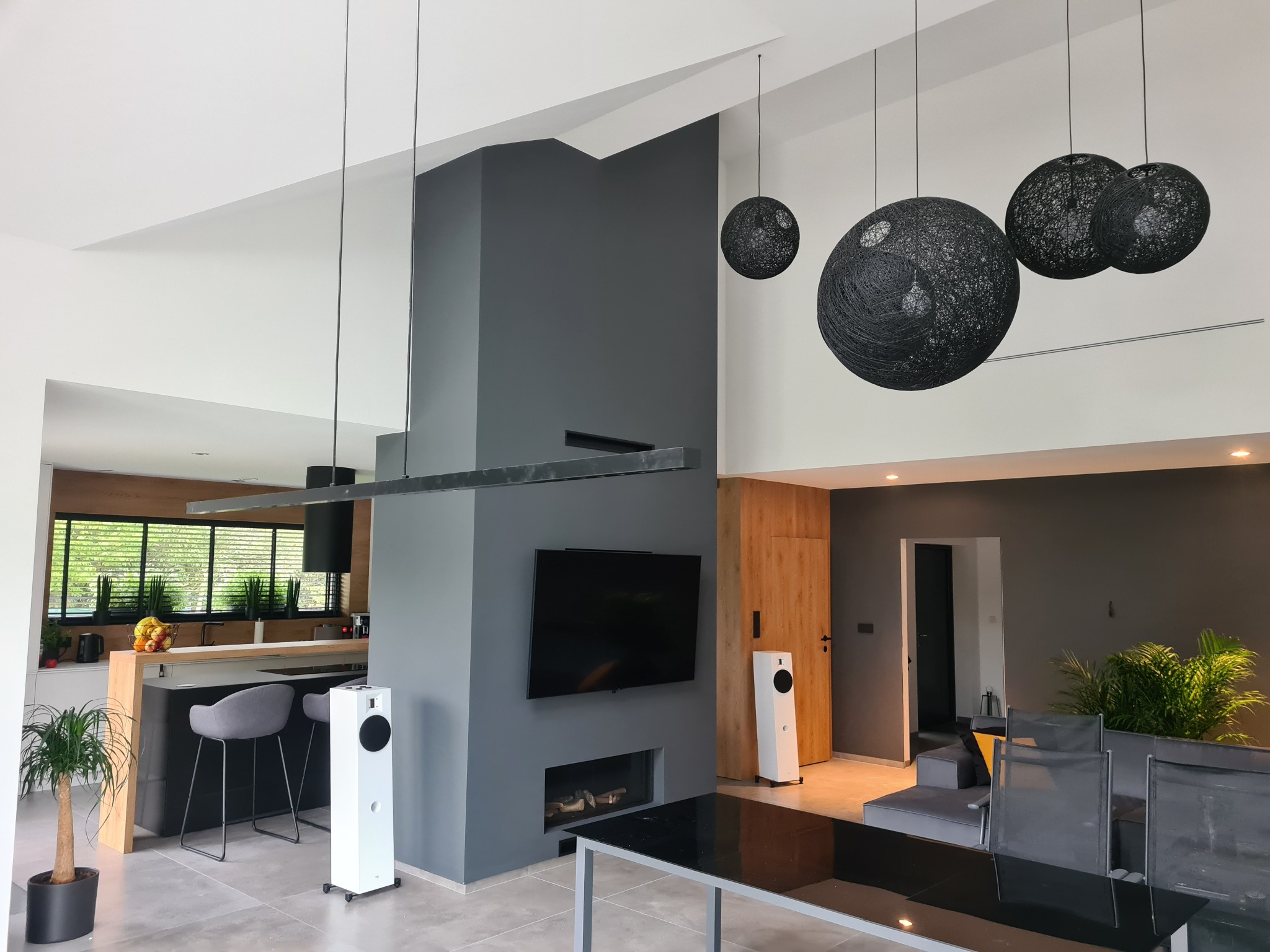 Wnętrze nowoczesnego salonu z aneksem kuchennym, szara ściana z telewizorem i kominkiem, wiszące czarne lampy kuliste, białe kolumny głośnikowe, stół z czarnym blatem, szare krzesła barowe przy...