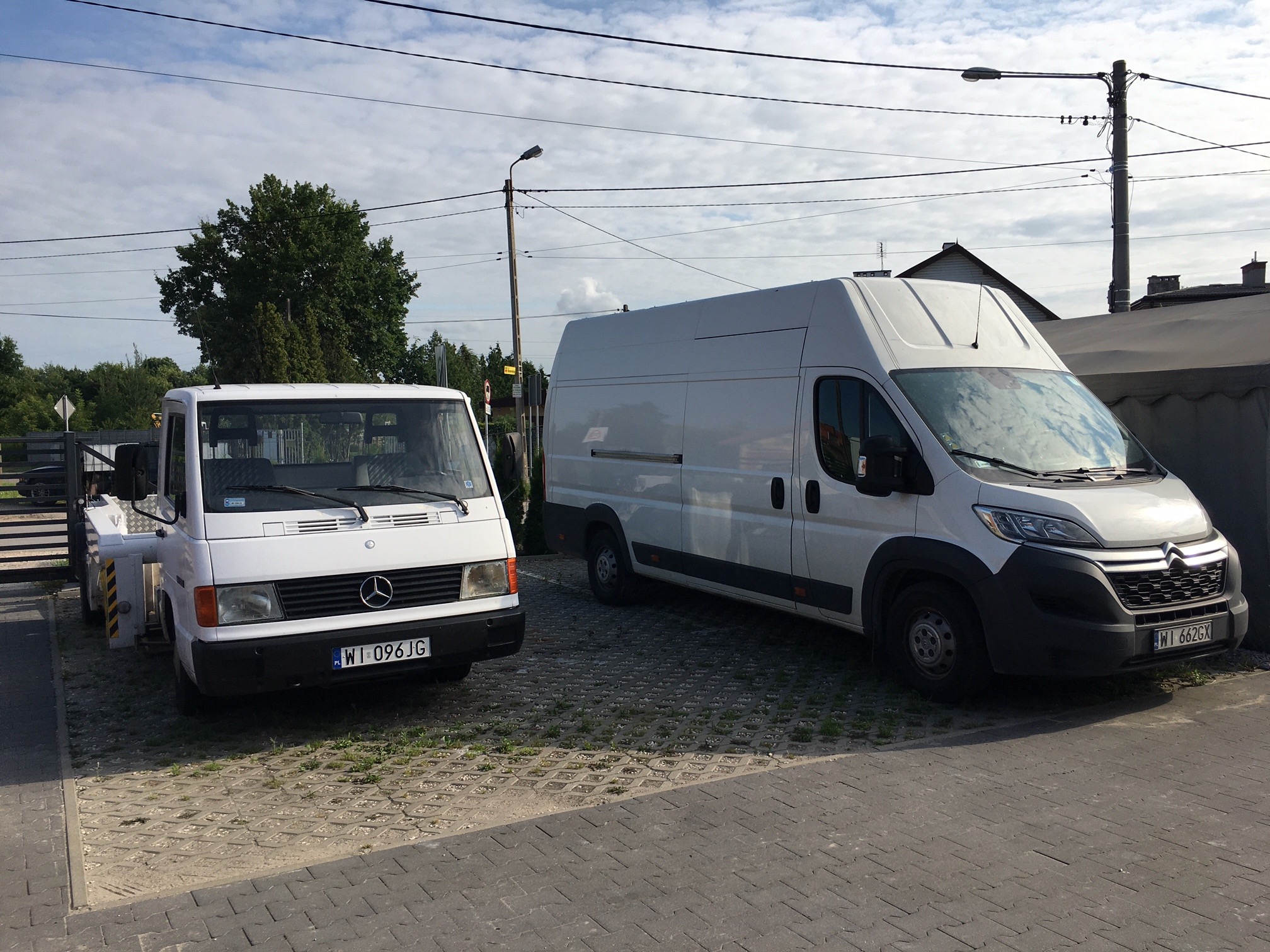 Dwa białe samochody dostawcze zaparkowane na kostce brukowej: starszy Mercedes-Benz laweta z podnośnikiem i nowszy Citroen Jumper
