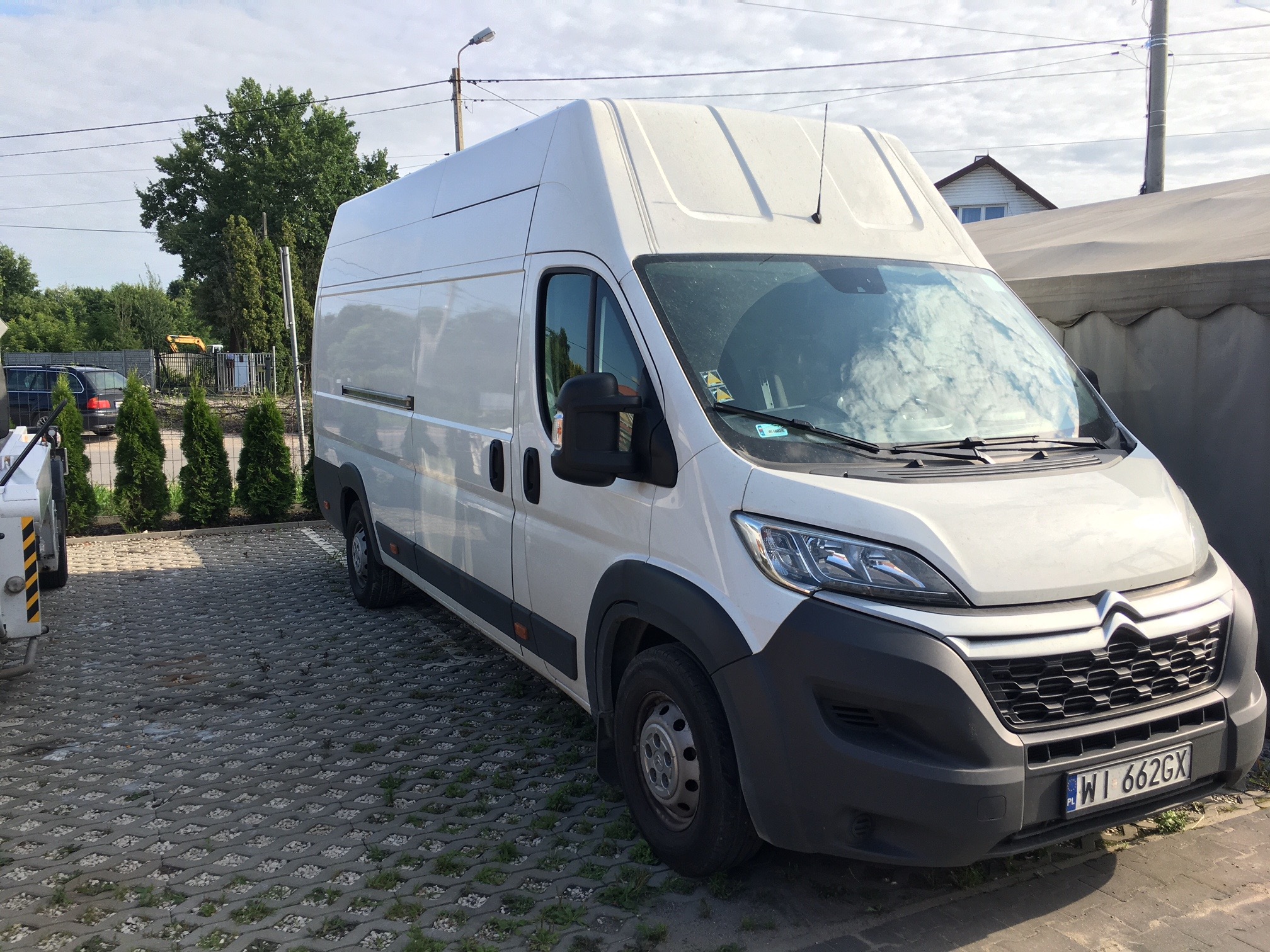 Biały, wysoki bus marki Citroen z widoczną tablicą rejestracyjną WI 662GX, zaparkowany na ażurowym parkingu z kostki betonowej, w tle zieleń i budynki.