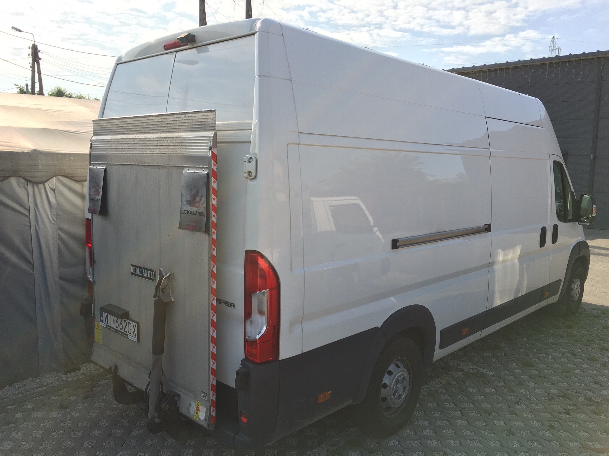 Biały bus dostawczy z otwartą windą załadowczą marki Dhollandia, zaparkowany na ażurowym parkingu, widok z tyłu i boku.