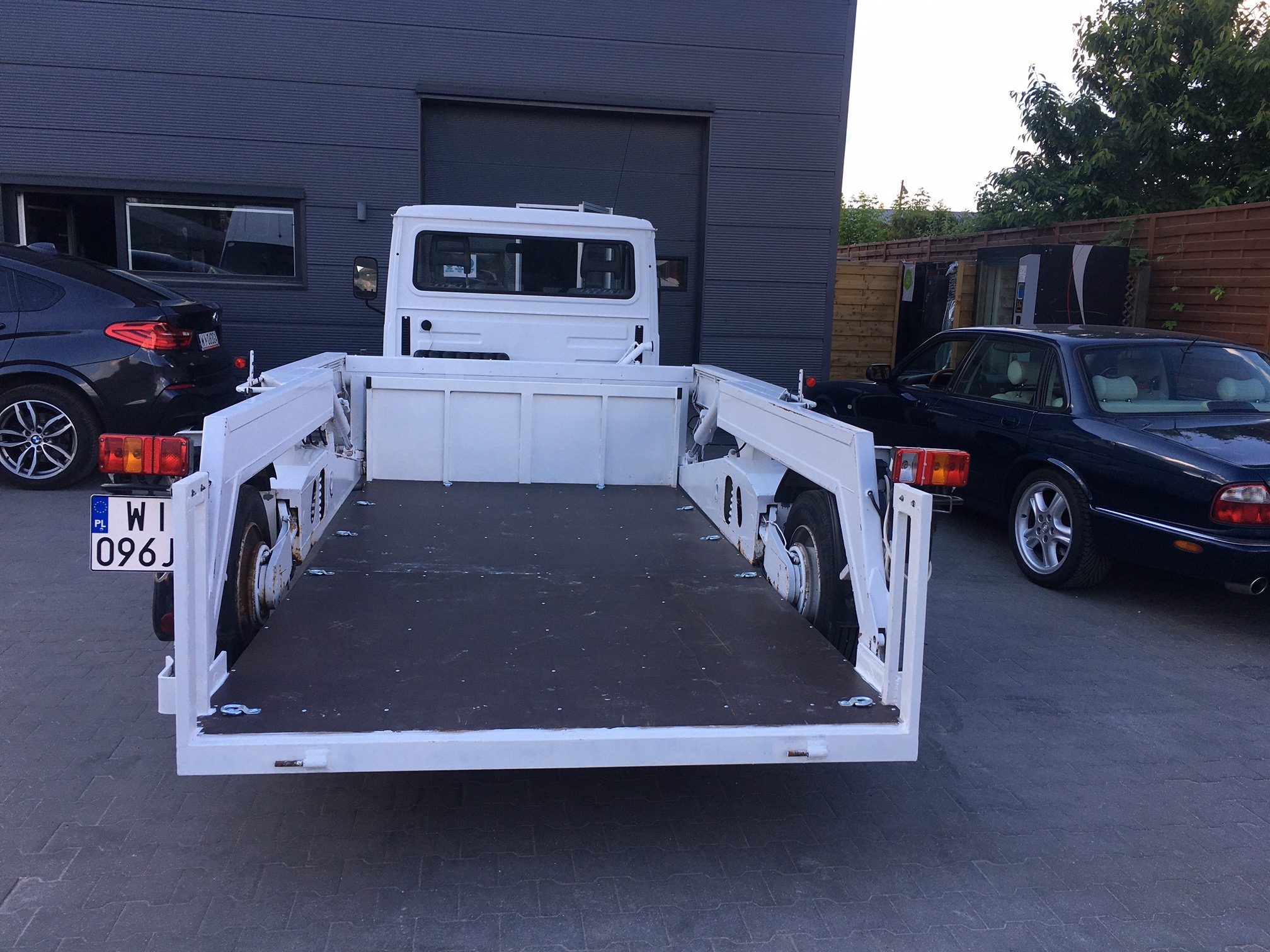 Biała laweta z opuszczanym najazdem, widok z tyłu, z zaparkowanymi samochodami osobowymi w tle.