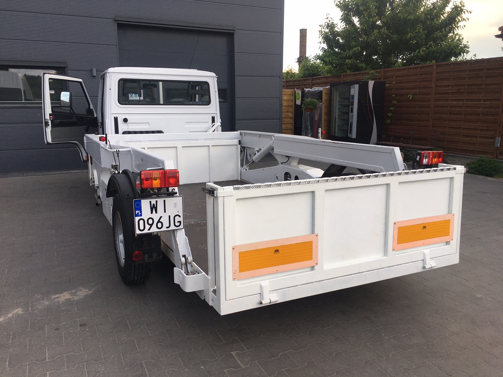 Biała, pusta platforma transportowa z otwartymi drzwiami kabiny, zaparkowana na szarym bruku przed szarą halą przemysłową, widoczny numer rejestracyjny.