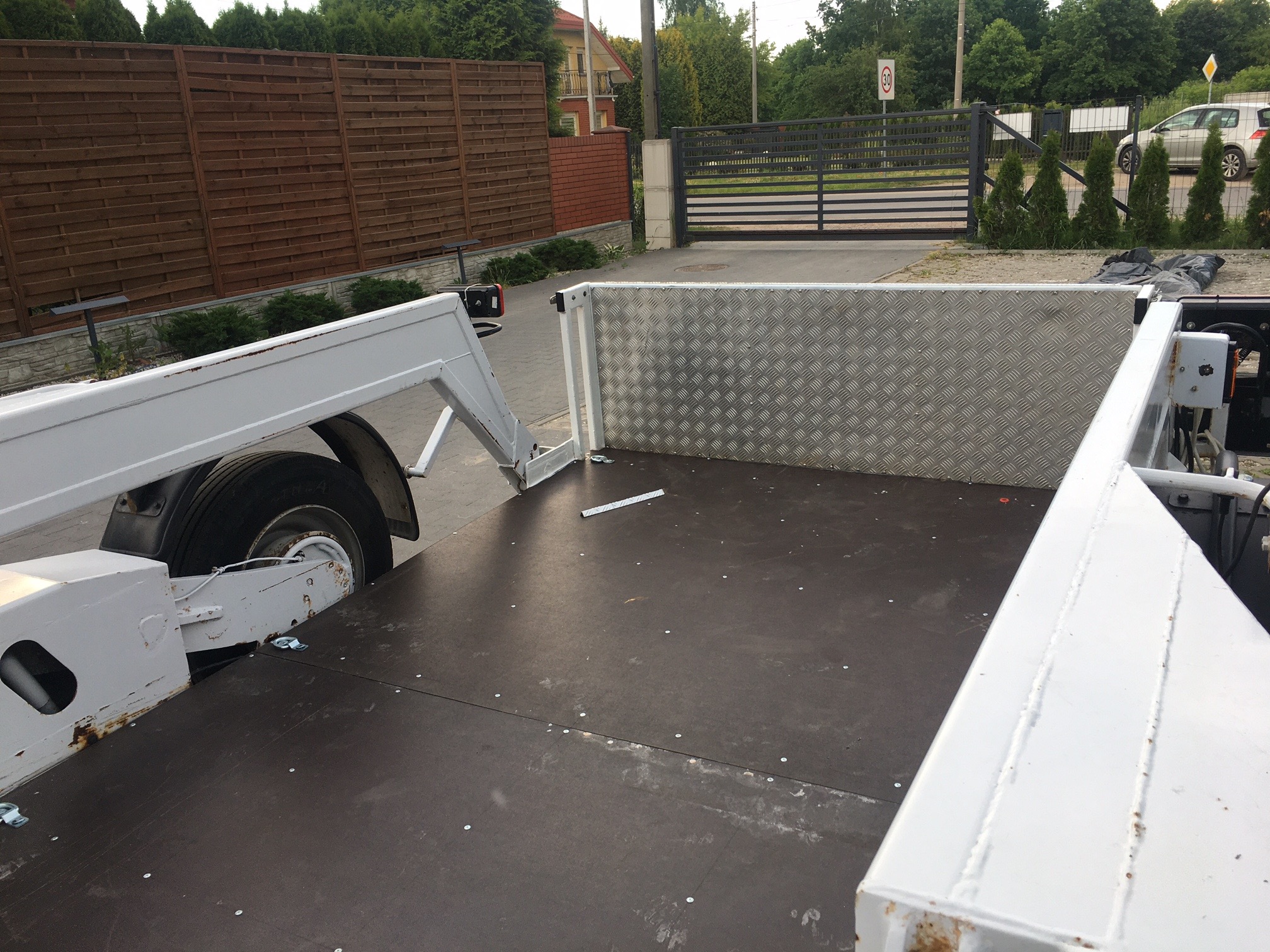 Pusta, biała laweta z podłogą z ciemnej sklejki, widok z wnętrza na zewnątrz, z widoczną konstrukcją i elementami mocującymi, na tle ogrodzenia posesji.