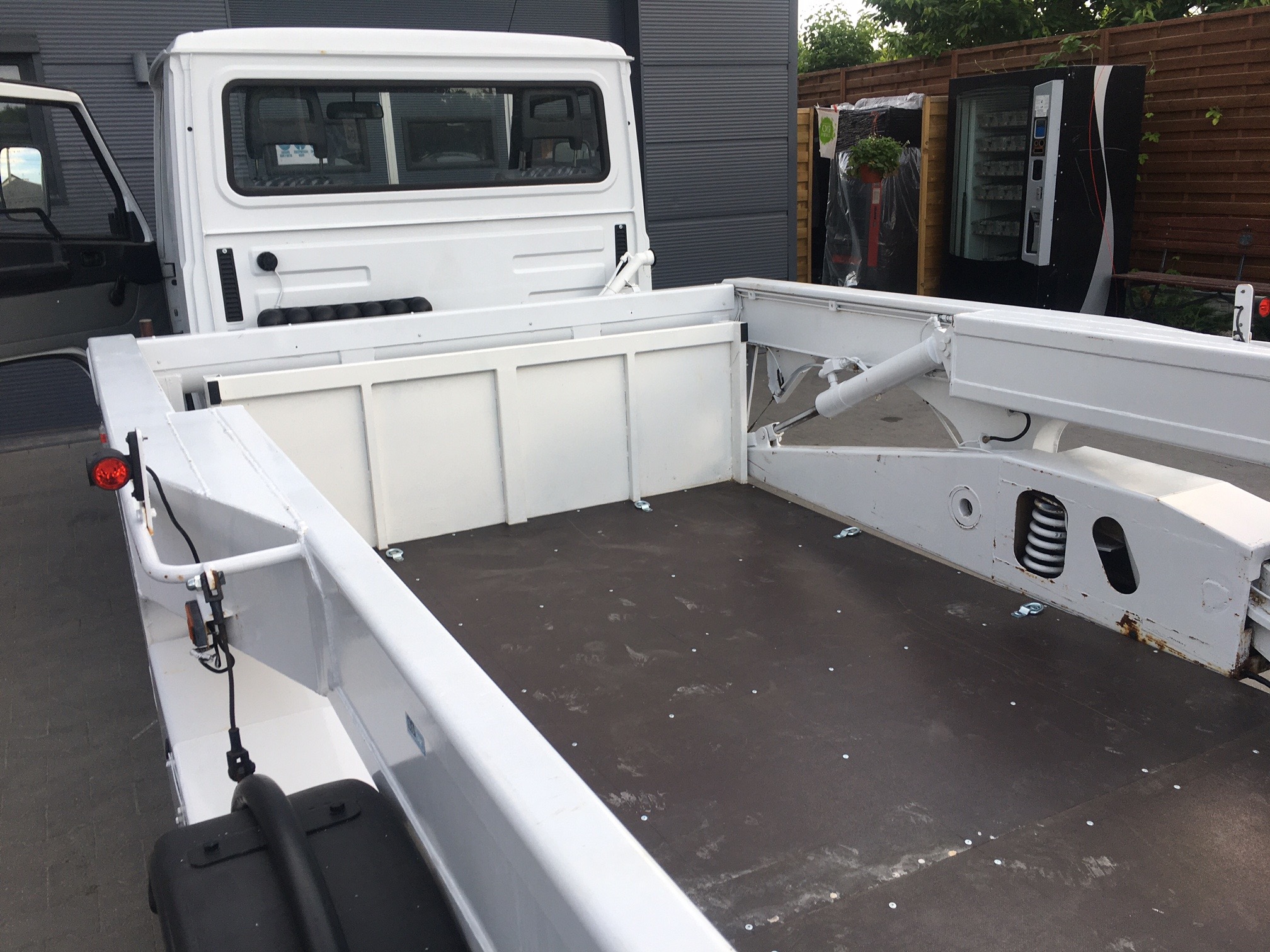 Pusta, biała platforma lawety z widocznymi elementami hydraulicznymi i mocowaniami, zaparkowana na zewnątrz z automatem do napojów w tle.