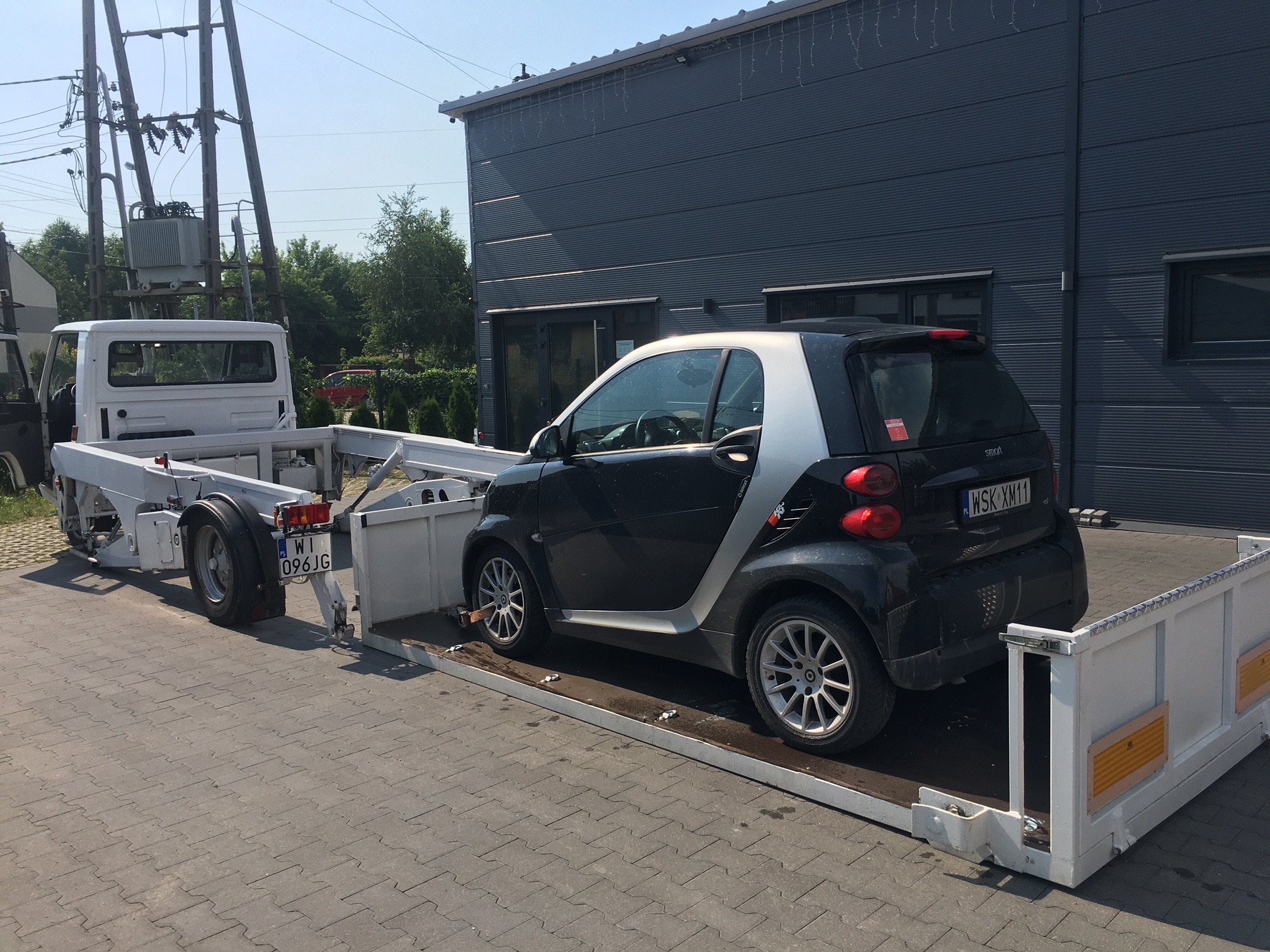 Czarny Smart załadowany na białą platformę lawety, widok z tyłu, budynek przemysłowy w tle.