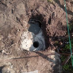 DEJV-BUD Cięcie i Wiercenie otworów w betonie, Technika Diamentowa - Wywiercony otwór w ziemi, częściowo wypełniony wodą, przygotowany pod fundament, widoczne ślady betonu i ziemi, obok fragment buta roboczego i element drewniany.