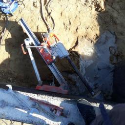 DEJV-BUD Cięcie i Wiercenie otworów w betonie, Technika Diamentowa - Wiertnica rdzeniowa z wiertłem w trakcie pracy przy fundamencie, obok leżą klucze płaskie i nastawny, widoczna woda w wykopie.