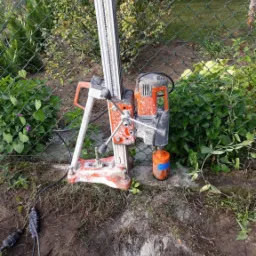 Wiertnica diamentowa z pomarańczowym silnikiem, ustawiona na betonowej powierzchni obok zielonego ogrodzenia z siatki, ślady wiercenia w ziemi.