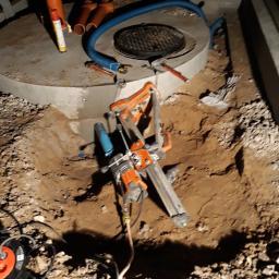 DEJV-BUD Cięcie i Wiercenie otworów w betonie, Technika Diamentowa - Betonowy krąg z włazem w ziemi, obok rury kanalizacyjne i wiertnica diamentowa do betonu z podłączonym przewodem zasilającym.