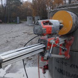 DEJV-BUD Cięcie i Wiercenie otworów w betonie, Technika Diamentowa - Wiertnica diamentowa Husqvarna mocowana do betonowego elementu, widoczne ślady wiercenia, w tle teren budowy z betonowymi kręgami.