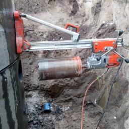 DEJV-BUD Cięcie i Wiercenie otworów w betonie, Technika Diamentowa - Wiertnica diamentowa w trakcie wiercenia otworu w betonowym szambie, widoczne zarysowania na powierzchni szamba i mokra ziemia wokół.