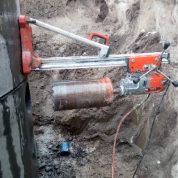 Wiertnica diamentowa w trakcie wiercenia otworu w betonowym szambie, widoczne zarysowania na powierzchni szamba i mokra ziemia wokół.