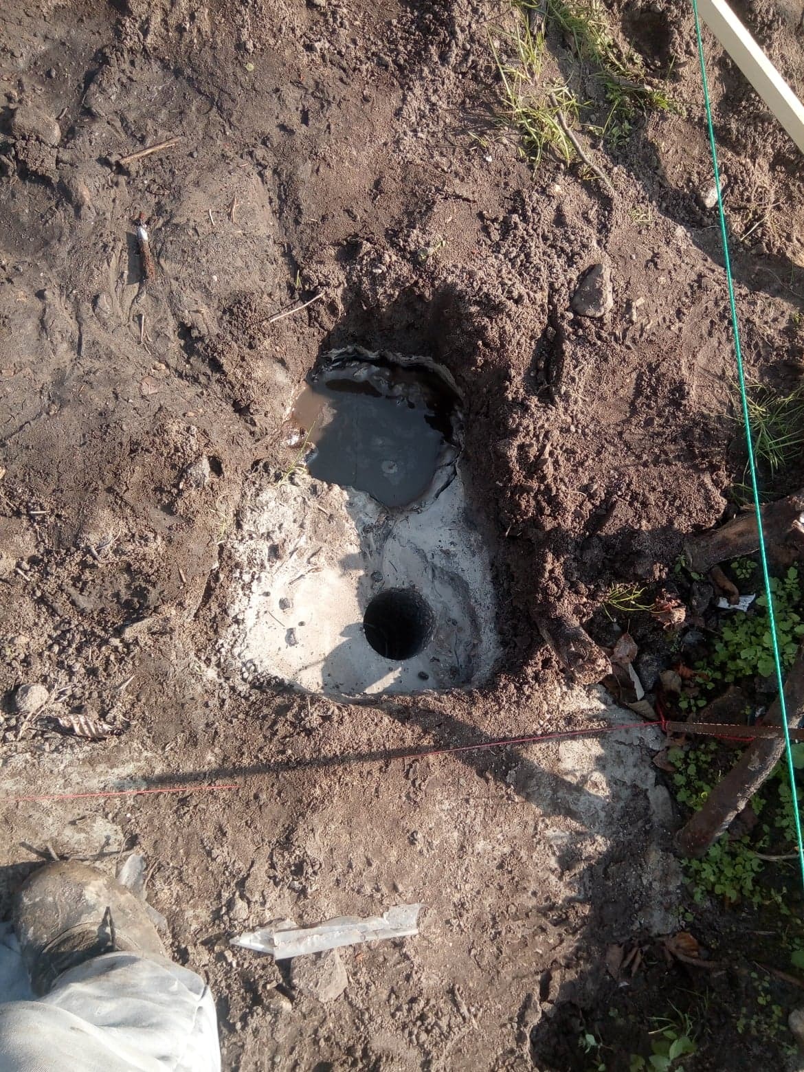 Wywiercony otwór w ziemi, częściowo wypełniony wodą, przygotowany pod fundament, widoczne ślady betonu i ziemi, obok fragment buta roboczego i element drewniany.