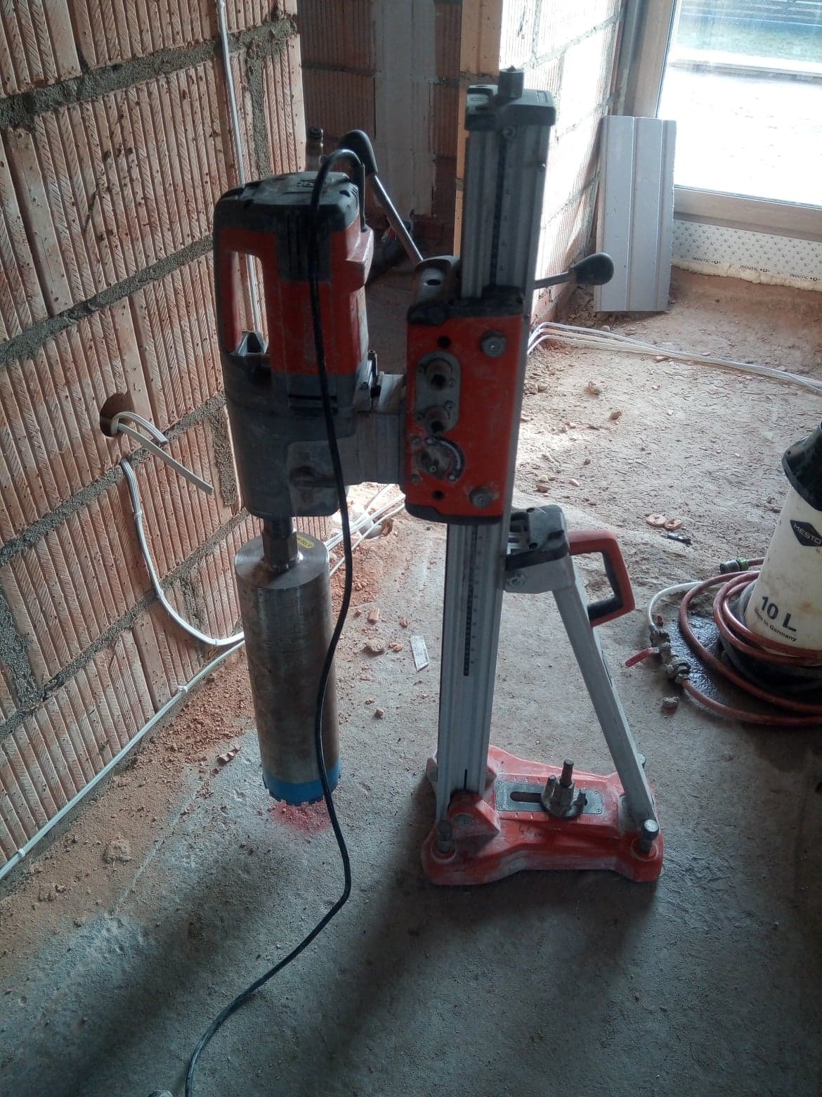 Wiertnica diamentowa z koroną w trakcie wiercenia otworu w ścianie z czerwonej cegły, widoczne przewody elektryczne i ślady wiercenia na podłodze.