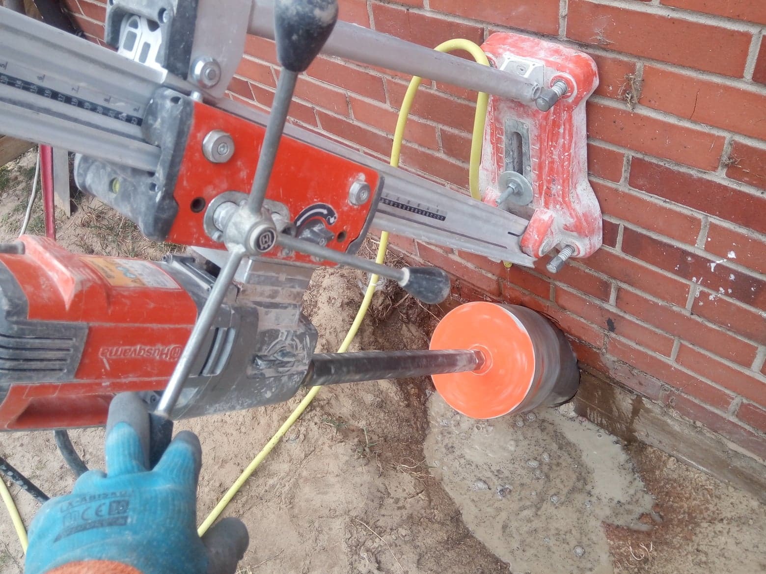 Wiercenie rdzeniowe w ceglanej ścianie z użyciem wiertnicy Husqvarna na mokro, widoczna podpora wiertnicy zamocowana do ściany, operator w niebieskiej rękawicy.