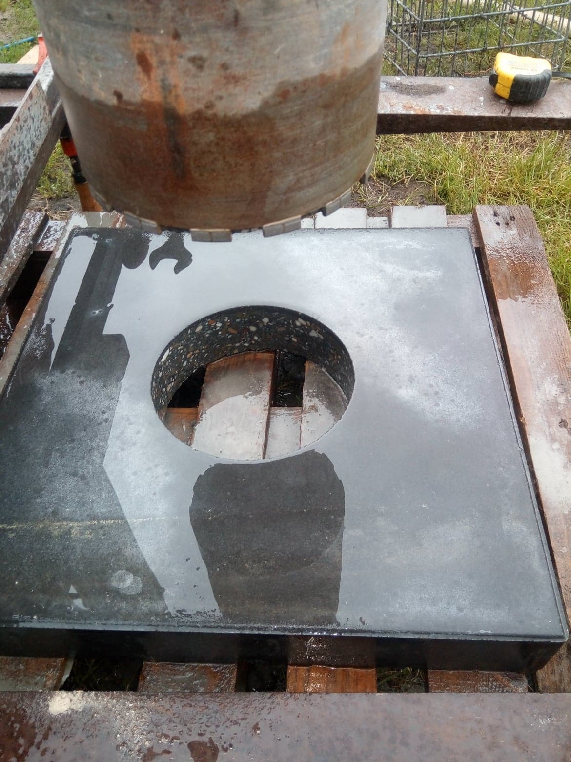 Wiertnica rdzeniowa z diamentową koronką nad mokrą płytą betonową z wywierconym otworem, widoczne ślady wody i błota.