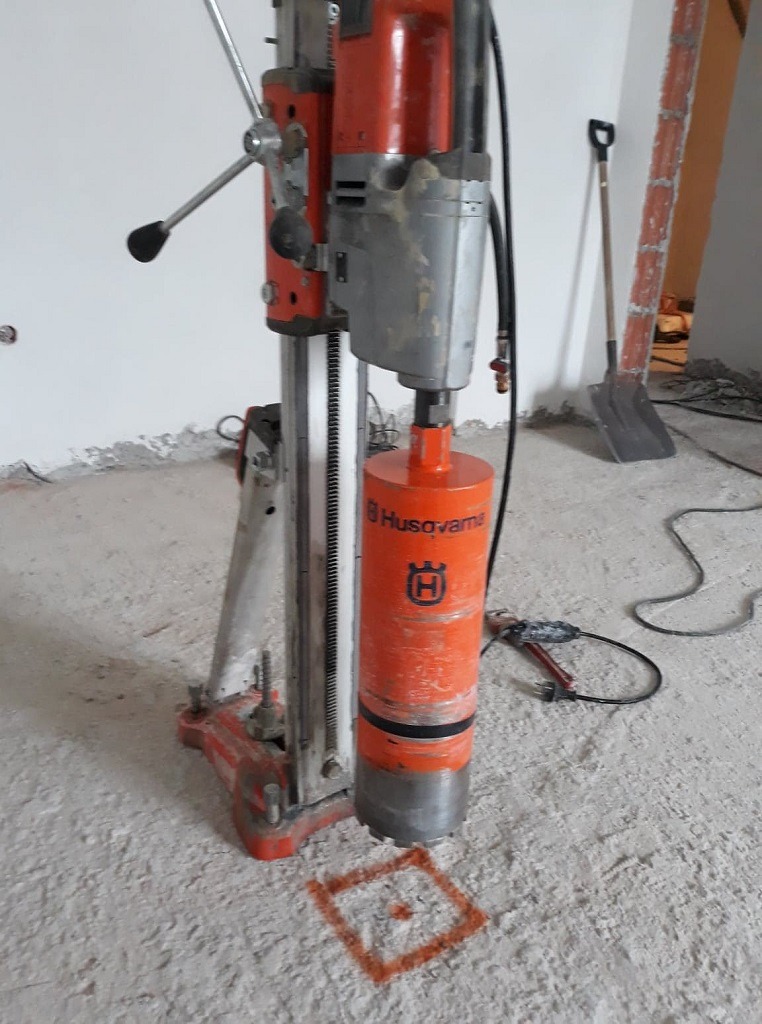 Wiertnica Husqvarna z wiertłem koronowym, przygotowana do wiercenia otworu w betonowej posadzce, zaznaczonego pomarańczowym kwadratem.