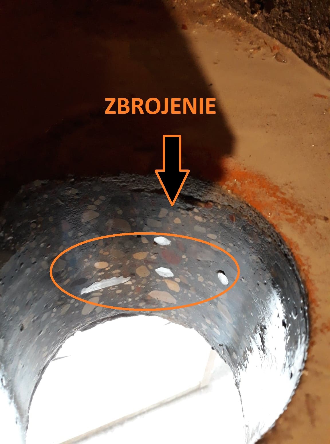 Wnętrze betonowego kręgu szamba z widocznym zbrojeniem i fragmentami białego tworzywa w strukturze betonu.