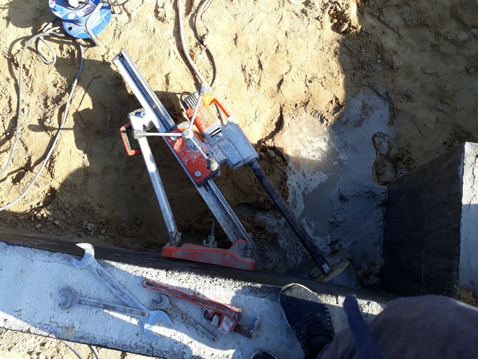 Wiertnica rdzeniowa z wiertłem w trakcie pracy przy fundamencie, obok leżą klucze płaskie i nastawny, widoczna woda w wykopie.