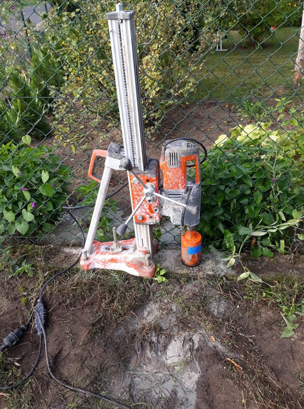 Wiertnica diamentowa z pomarańczowym silnikiem, ustawiona na betonowej powierzchni obok zielonego ogrodzenia z siatki, ślady wiercenia w ziemi.