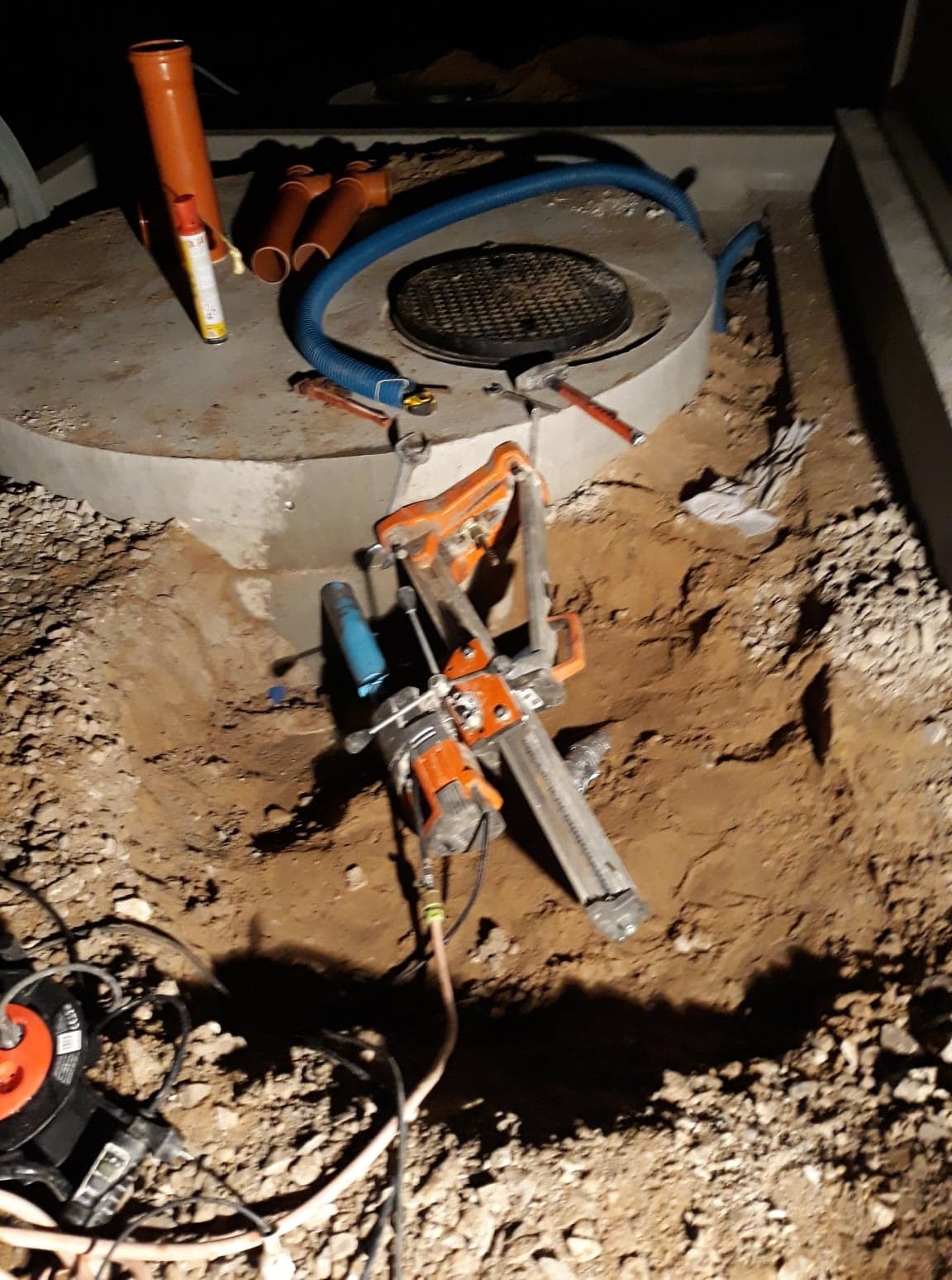 Betonowy krąg z włazem w ziemi, obok rury kanalizacyjne i wiertnica diamentowa do betonu z podłączonym przewodem zasilającym.