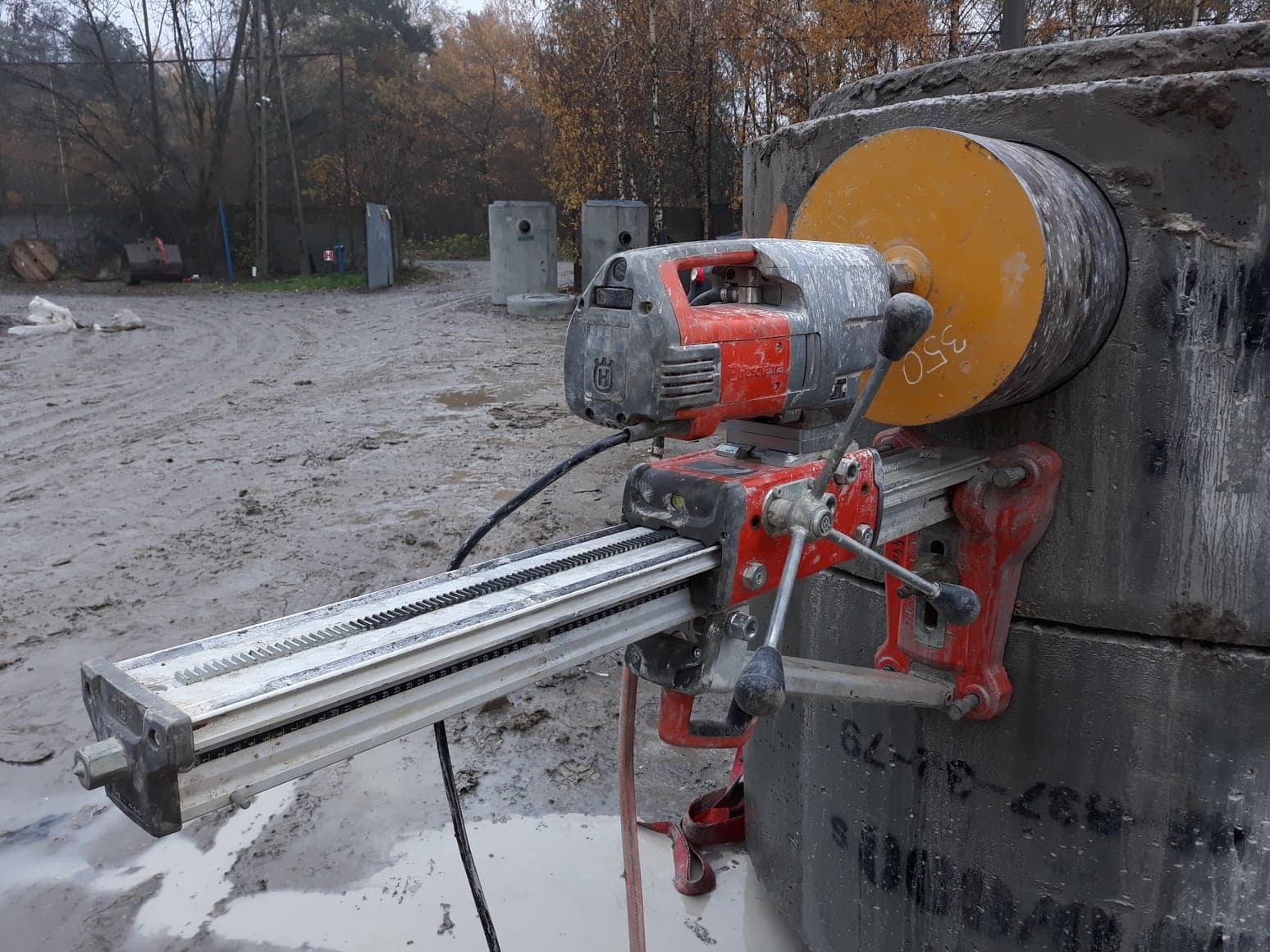 Wiertnica diamentowa Husqvarna mocowana do betonowego elementu, widoczne ślady wiercenia, w tle teren budowy z betonowymi kręgami.