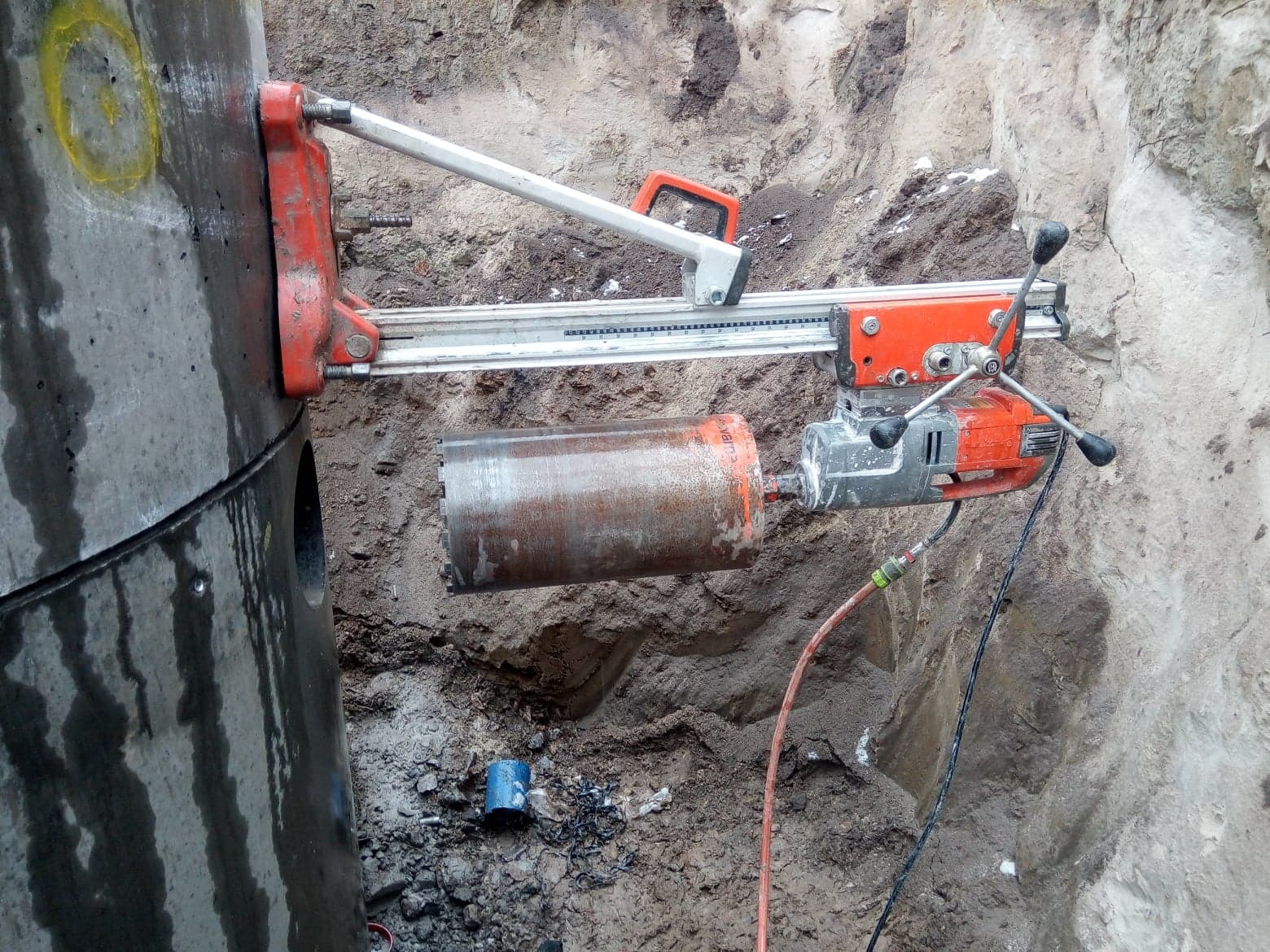 Wiertnica diamentowa w trakcie wiercenia otworu w betonowym szambie, widoczne zarysowania na powierzchni szamba i mokra ziemia wokół.