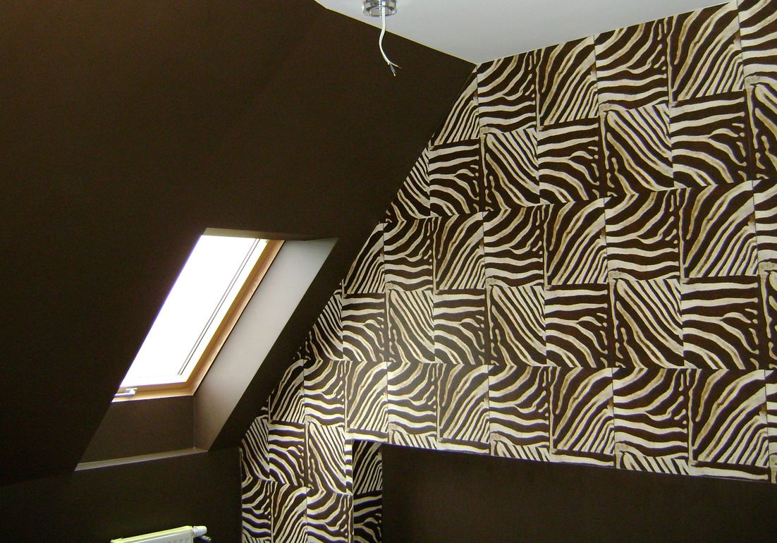 Pokój na poddaszu z oknem dachowym i ścianą pokrytą tapetą w motywy zebry, brązowa ściana, widoczne kable od lampy sufitowej.