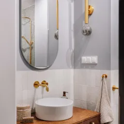 Nowoczesna łazienka z okrągłą białą umywalką nablatową, drewnianą szafką, złotą armaturą, owalnym lustrem i designerską lampą ścienną z szarymi żarówkami.