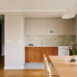 Jasna kuchnia z drewnianymi szafkami dolnymi i beżowymi górnymi, marmurowym backsplashiem, drewnianym parkietem w jodełkę i stołem z kwiatami w wazonie.