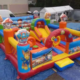 Kolorowy dmuchany plac zabaw z motywem bajki 'Psi Patrol' na niebieskiej plandece, z widocznym białym namiotem i budynkami w tle.