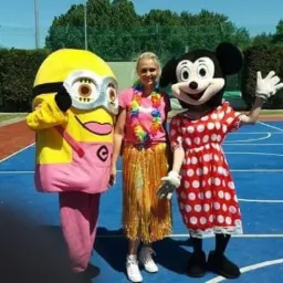 Trzy osoby w kostiumach: Minionka, kobiety w hawajskim stroju i Myszki Miki, stojące na niebieskim boisku sportowym.