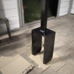 Usługi spawalnicze Dawid Kruk - Czarny, metalowy słupek z podstawą w kształcie litery U, stojący na kartonie na tle kostki brukowej i drzwi wejściowych.