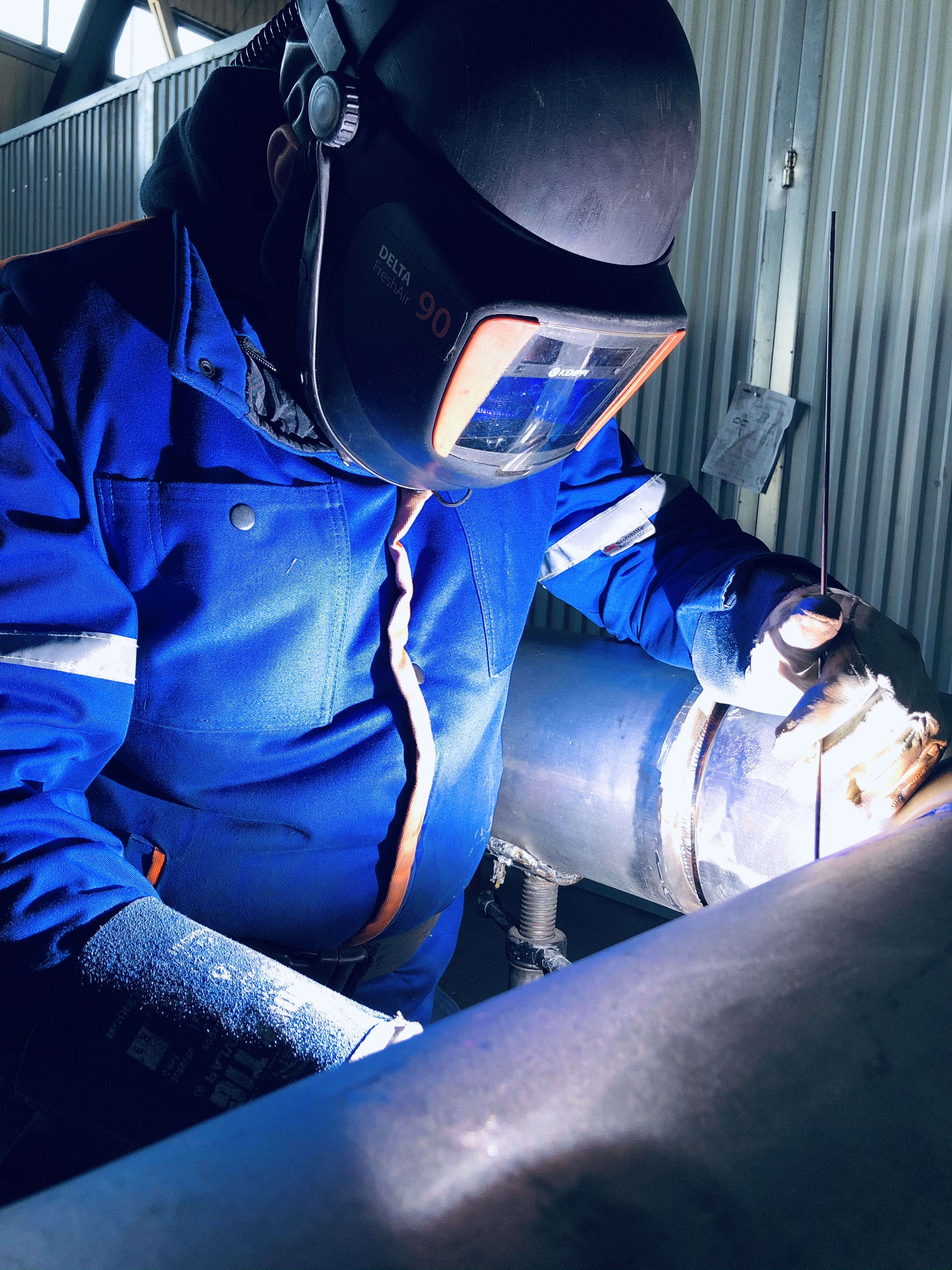 Pracownik w niebieskim kombinezonie i masce spawalniczej Delta FreshAir 90 łączy metalową rurę w pionowej pozycji, widoczne iskry i elektroda spawalnicza.