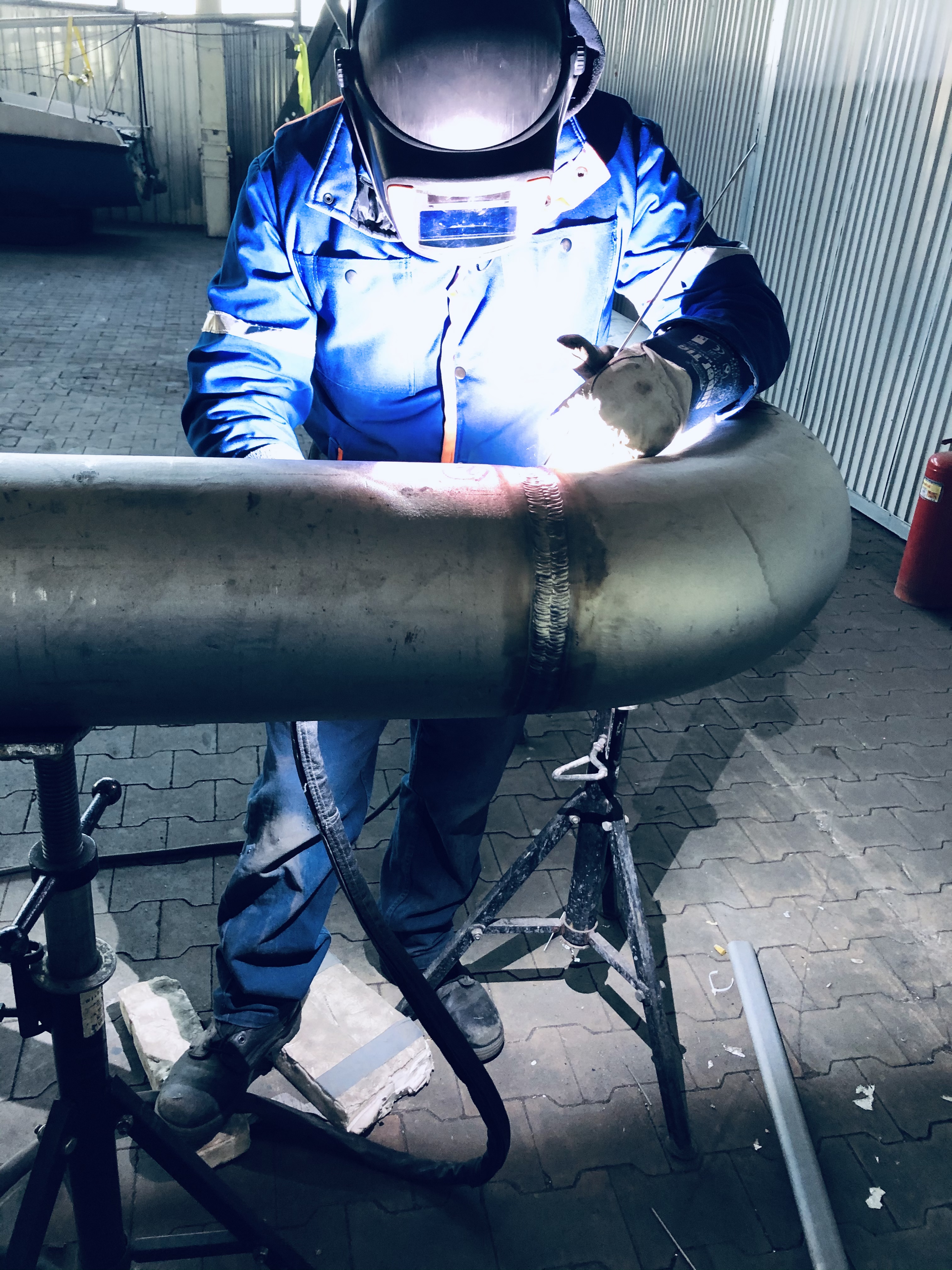 Pracownik w niebieskim kombinezonie i masce spawalniczej łączy spawarką dwa elementy rury na stojaku, widoczne ślady spawania, w tle ściana z blachy falistej i gaśnica.