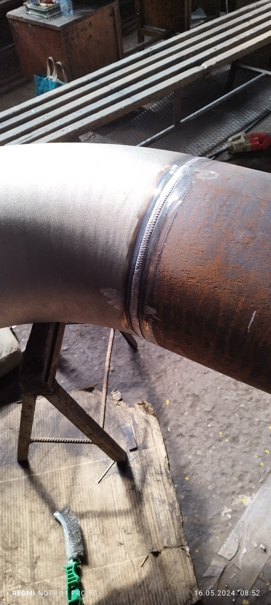 Zespawany kolanko rury, widoczny spaw na styku dwóch elementów, podparte na metalowym stojaku, obok leży szczotka druciana.