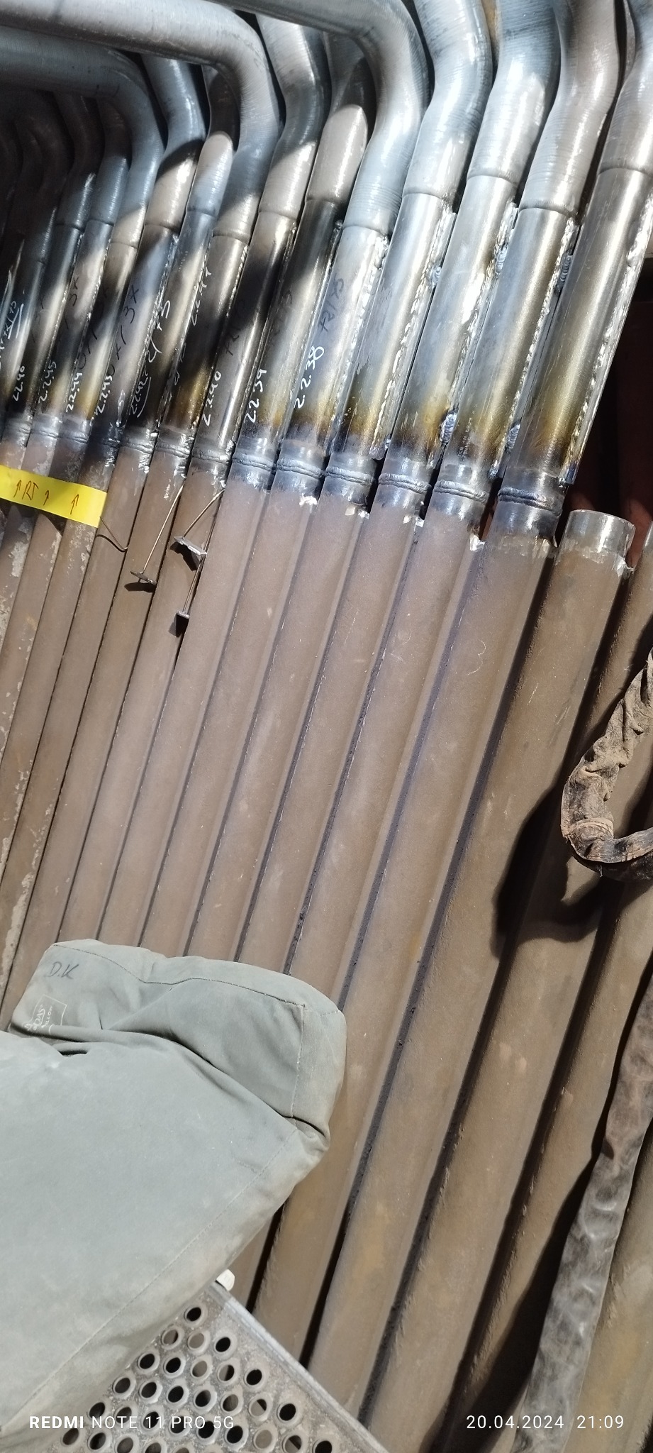 Rząd metalowych rur połączonych spawami, z widocznymi numerami i oznaczeniami, leżących ukośnie na szarej powierzchni, z fragmentem ubrania roboczego w lewym dolnym rogu.