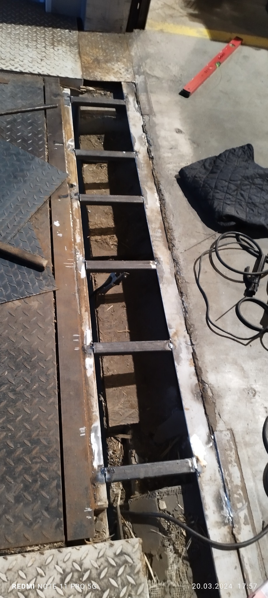 Spawana metalowa drabinka w trakcie montażu w podłodze, widoczne ślady spawania, poziomica i przewody w tle.