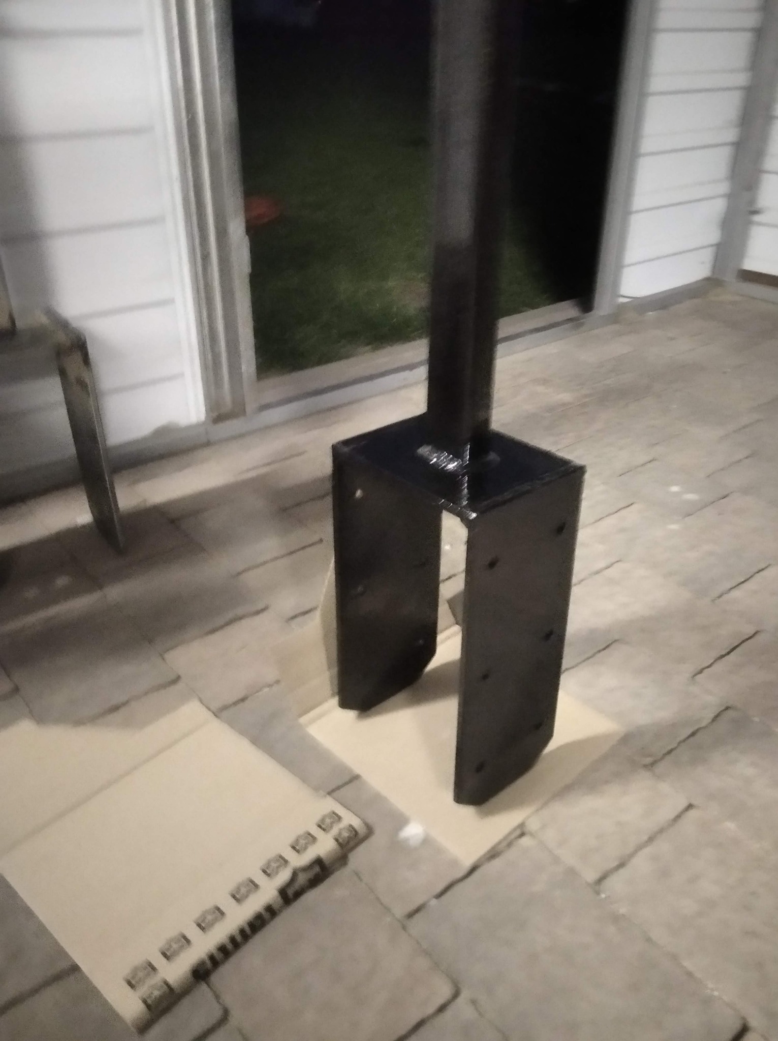 Czarny, metalowy słupek z podstawą w kształcie litery U, stojący na kartonie na tle kostki brukowej i drzwi wejściowych.