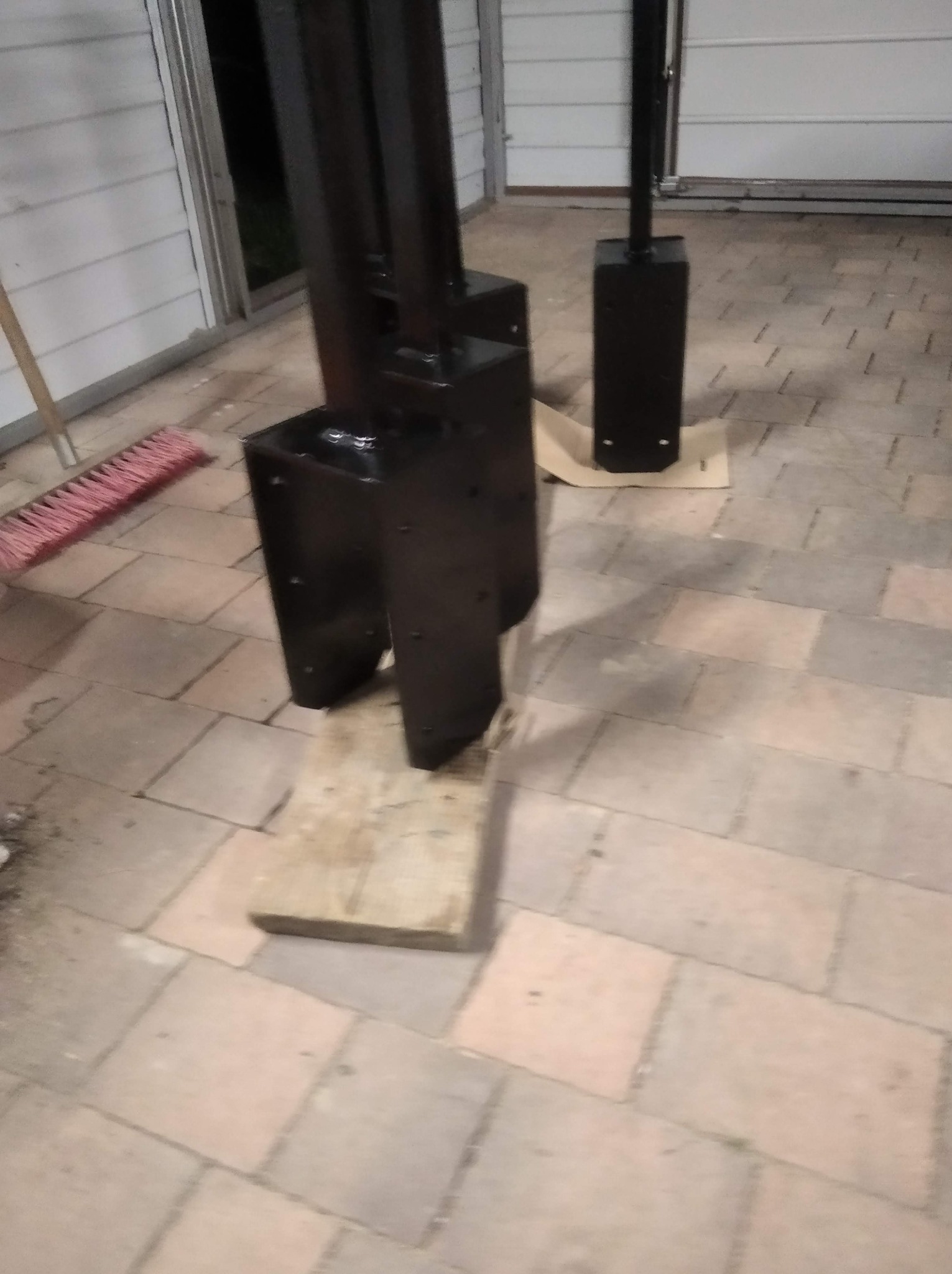 Czarne, metalowe konstrukcje po obróbce, stojące na drewnianych podkładkach na tle garażu z kostką brukową i miotłą.