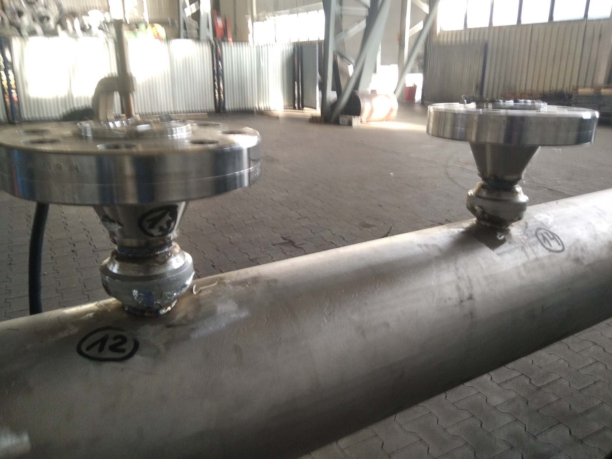 Spawane połączenie kołnierzowe rury stalowej z oznaczeniem numeru '12' i '14', widoczne ślady spawania i obróbki, tło hali przemysłowej.