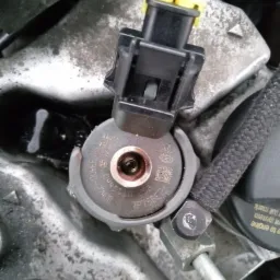 Zbliżenie na wtryskiwacz paliwa z żółtym złączem elektrycznym, widoczne numery seryjne i kod QR na szarej obudowie, element w silniku samochodowym.