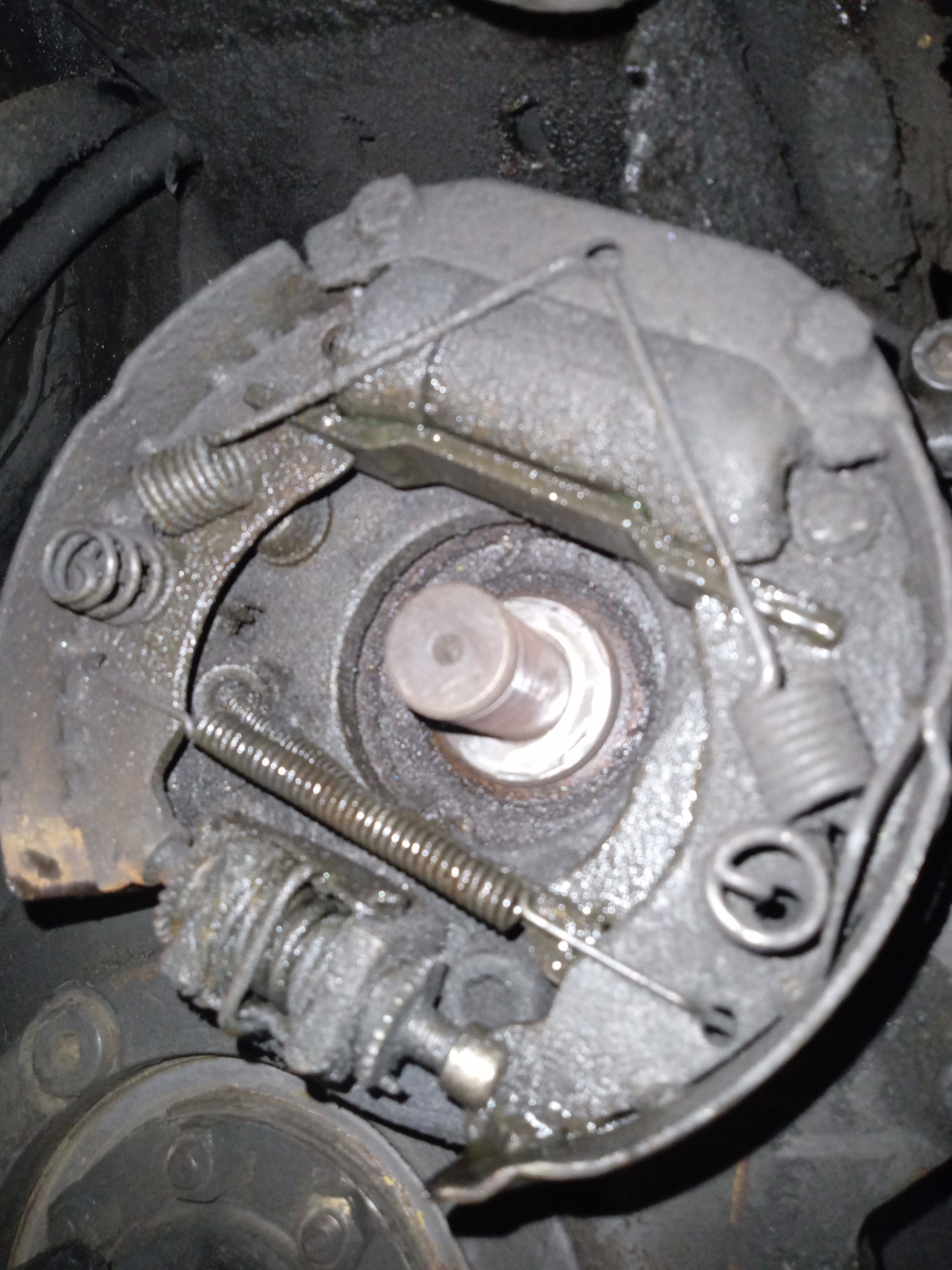 Zużyty i brudny mechanizm hamulcowy bębnowy z widocznymi sprężynami, linkami i elementami metalowymi, pokryty smarem i brudem.