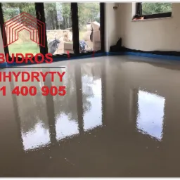 Świeżo wylana, gładka i lśniąca posadzka w jasnym pomieszczeniu z dużym oknem tarasowym, zabezpieczona niebieską taśmą przy ścianach.