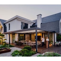 Pracownia Architektury Krajobrazu Błażej Niedbał - Nowoczesny ogród z drewnianym tarasem, pergolą i oświetleniem, idealny do relaksu. Para z kieliszkami wina dodaje scenie życia i elegancji. Kameralna atmosfera.