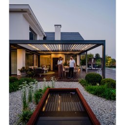 Pracownia Architektury Krajobrazu Błażej Niedbał - Nowoczesny ogród z pergolą i oczkiem wodnym, oświetlony wieczornym światłem. Para relaksuje się na tarasie z drewnianą podłogą i meblami ogrodowymi.