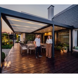 Pracownia Architektury Krajobrazu Błażej Niedbał - Nowoczesna pergola z regulowanym zadaszeniem nad drewnianym tarasem, oświetlony ogród w tle, para stojąca na tarasie, wieczorny krajobraz.