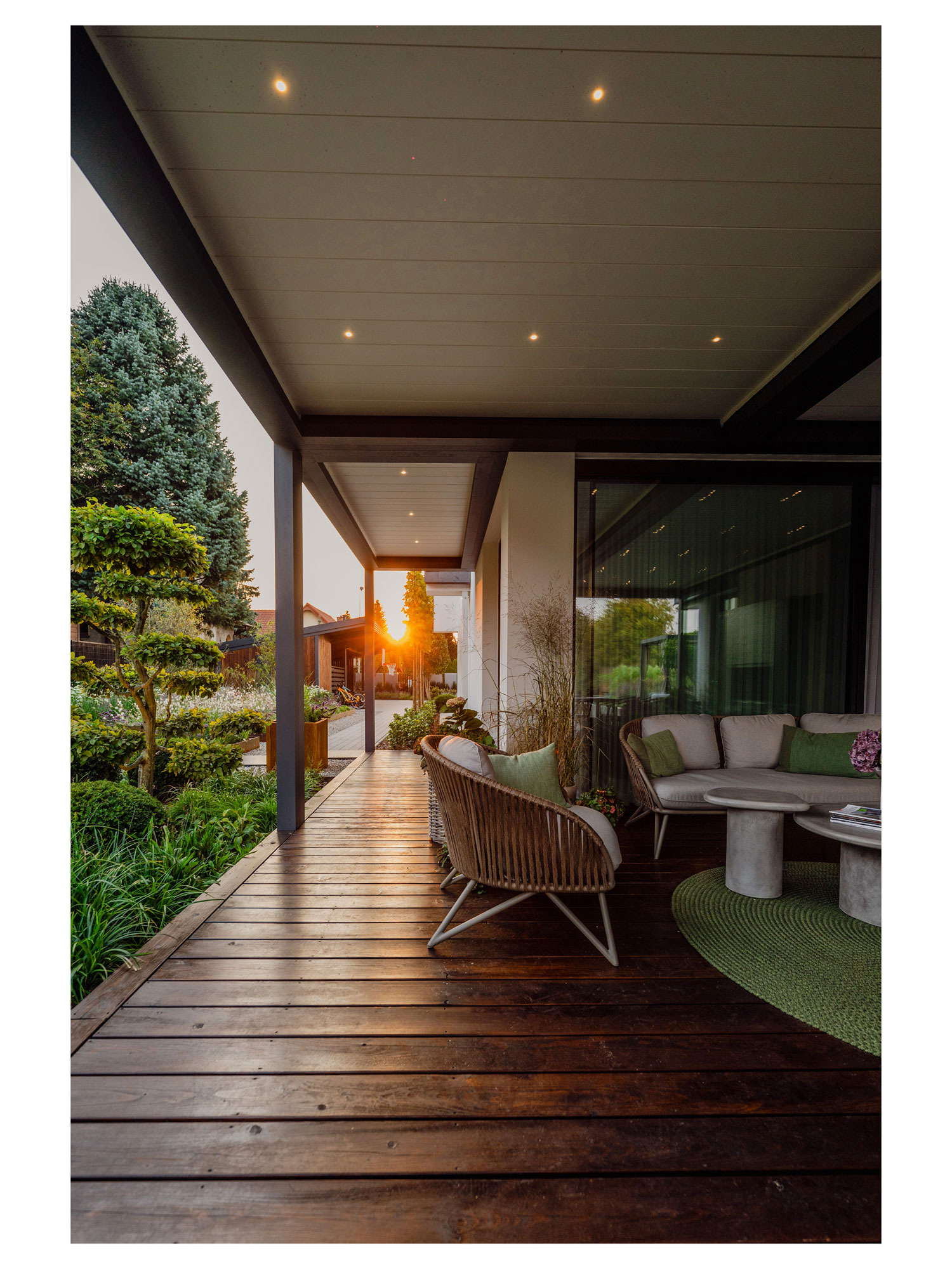 Zadaszony taras z drewnianą podłogą i meblami ogrodowymi w stylu boho, z widokiem na zadbany ogród o zachodzie słońca. Klimatyczne oświetlenie punktowe.