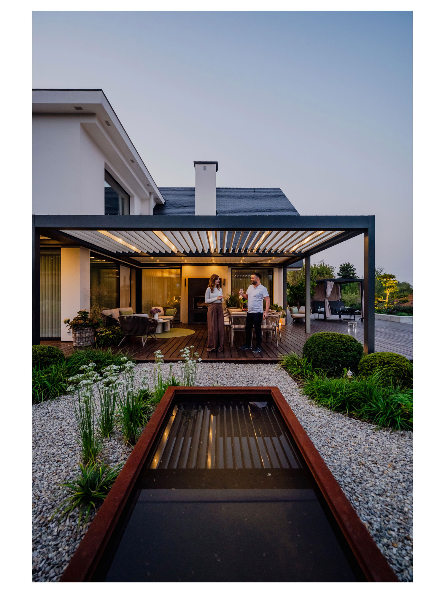 Nowoczesny ogród z pergolą i oczkiem wodnym, oświetlony wieczornym światłem. Para relaksuje się na tarasie z drewnianą podłogą i meblami ogrodowymi.